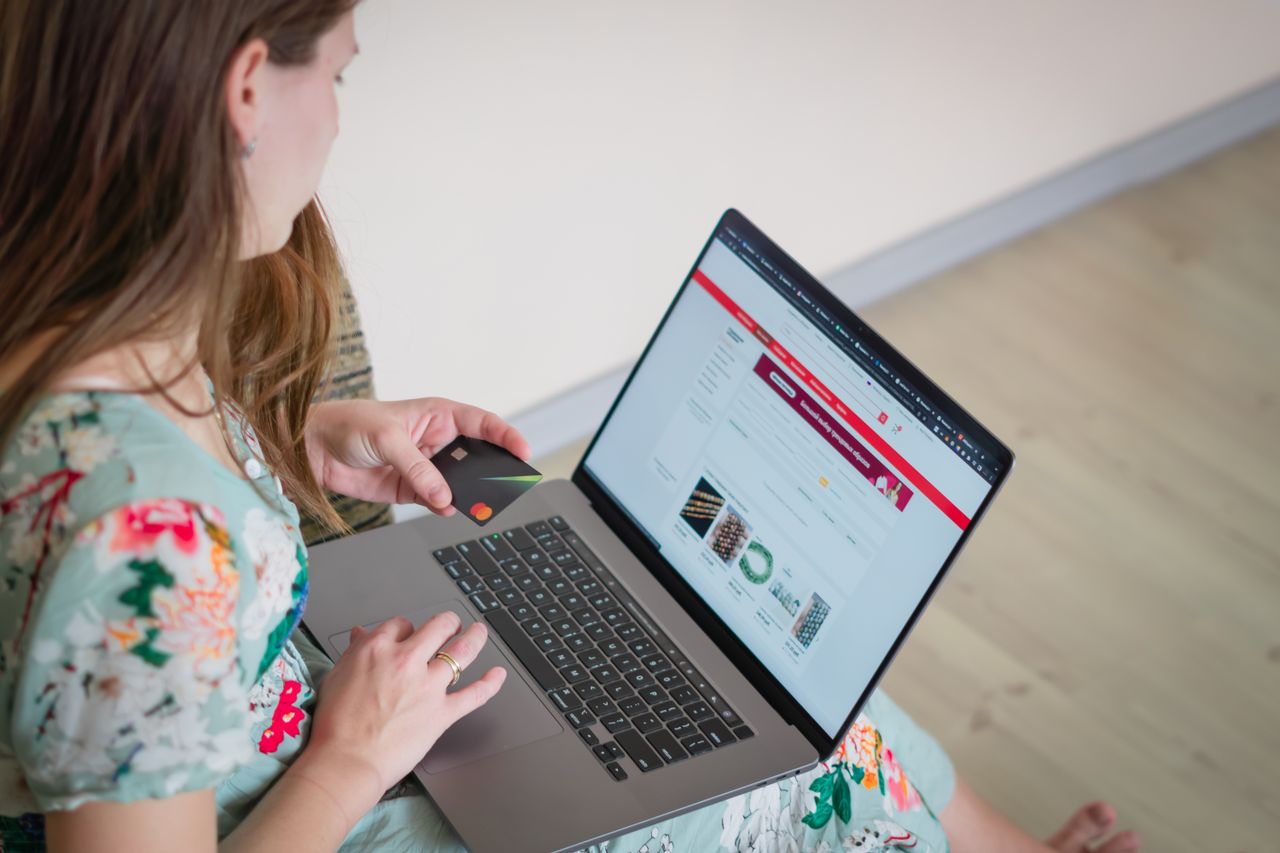 A woman wearing a floral dress is shopping online indoors with a laptop and a credit card.